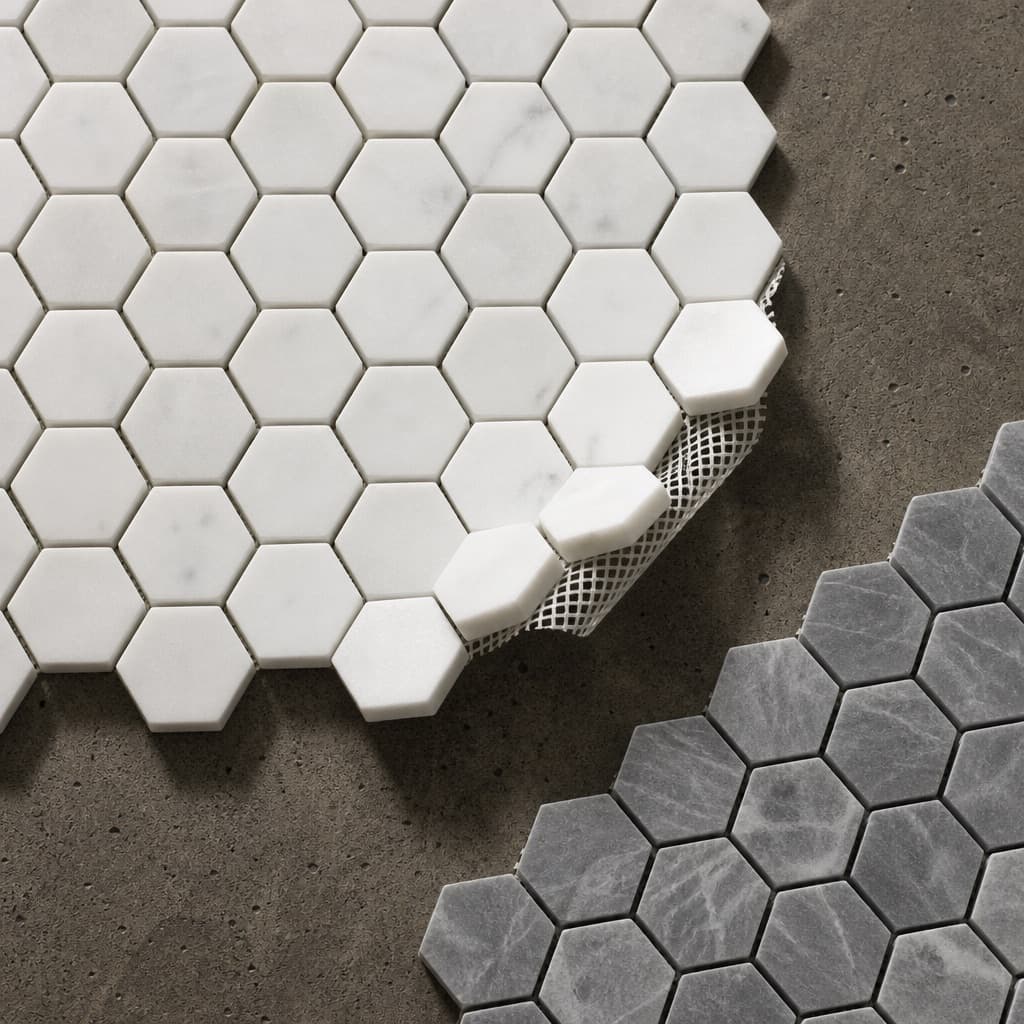 A sheet of white marble hexagon mosaic with one corner lifted to show the mesh backing, beside the corner of a grey bardiglio sheet